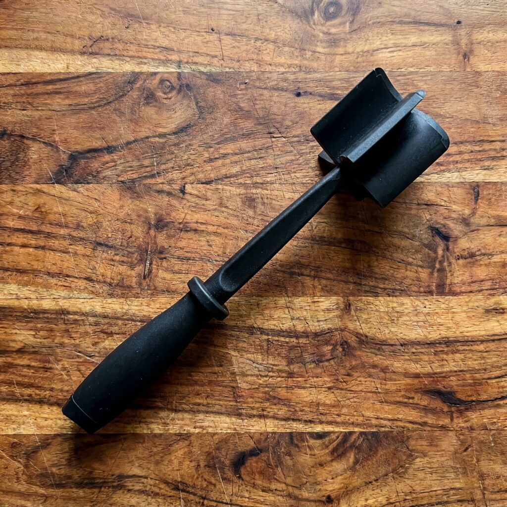 A black nylon meat chopper breaking up ground beef evenly in a frying pan.